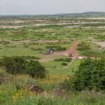 Utsikt från Observation Hill över en grön slätt med saltfläckar, en röd väg, tre 4x4-bilar parkerade vid en liten ockrafärgad byggnad, låga kullar i fjärran