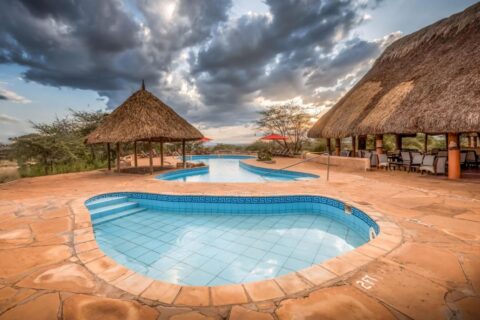 En liten kaklad bassäng i förgrunden och den stora poolen på Samburu Sopa Lodge, mellan en halmhydda och en terrass med halmtak
